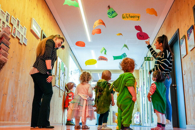 Kinder in Kostümen entdecken mit Erzieherinnen bunte Sprachschilder an der Decke.