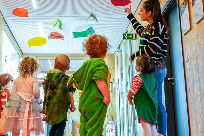 Kinder in Kostümen entdecken mit Erzieherinnen bunte Sprachschilder an der Decke.