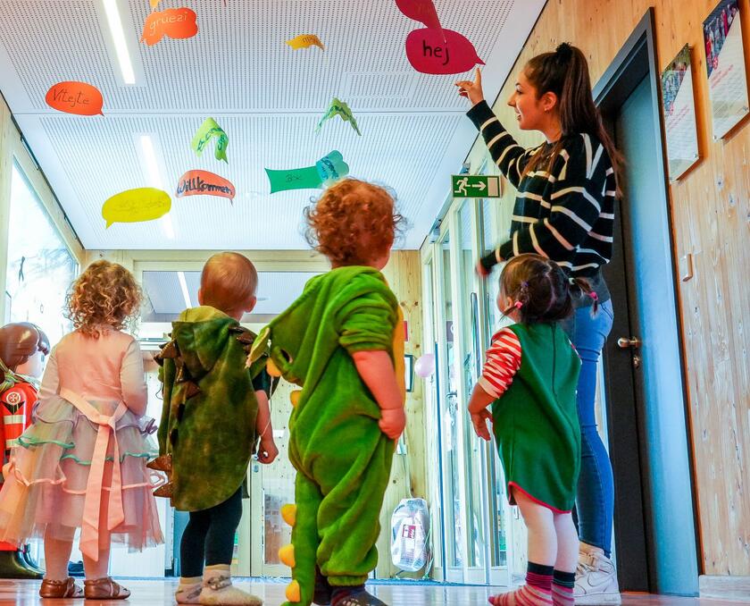 Kinder in Kostümen entdecken mit Erzieherinnen bunte Sprachschilder an der Decke.