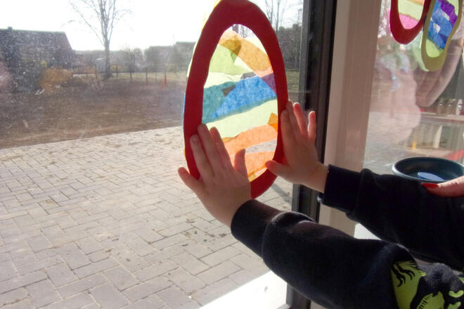 Ein Kind befestigt gerade ein großes selbstgebasteltes Osterei an der Fensterscheibe im Gruppenraum der Kita. 