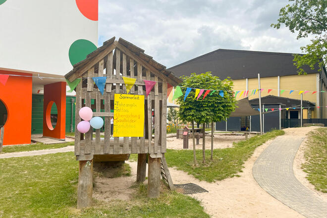 Eine Kinderholzhütte auf Stelzen mit einem gelben Pappschild auf dem "Sommerfest" steht. Dahinter einer Kita.