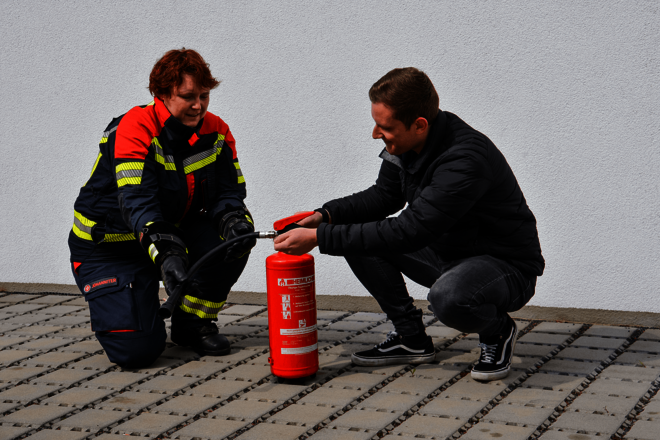 Ein Frau in Einsatzkleidung und ein Mann in dunkler Privatkleidung knieen auf Setinplatten neben einem Feuerlösche rund sprechen darüber. 