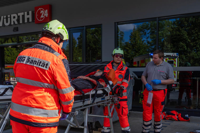 Zwei Ehrenamtliche Johanniter bringen eine verletzte Person auf einer Trage zum Behandlungsplatz und werden von einem weiteren Ehrenamtlichen eingewiesen.