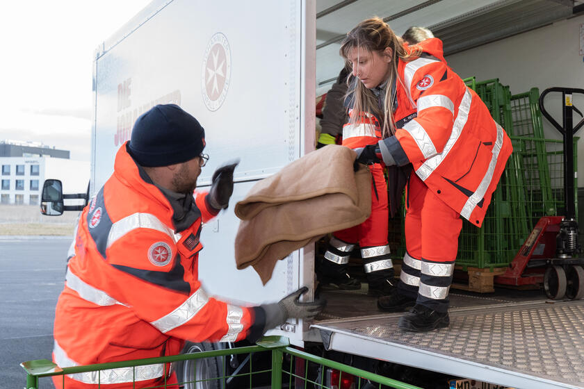 eine Mitarbeiterin der Johanniter Unfall Hilfe reicht einem anderen Mitarbeiter eine Decke aus dem Transporter