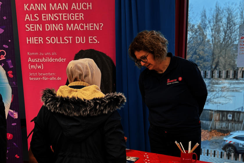 Eine Johanniter-Mitarbeiterin spricht auf einer Berufsmesse mit einer jungen Frau an einem Infostand. Im Hintergrund steht ein pinkes Roll-Up mit Informationen zur Ausbildung bei den Johannitern.