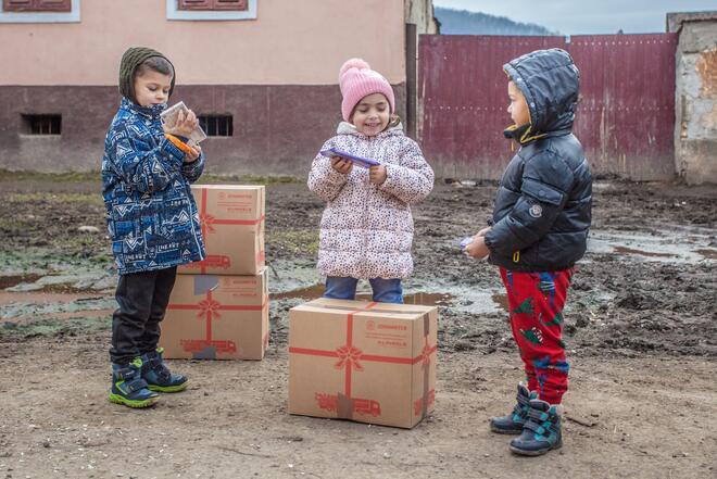 Drei kleine Kinder in Winterjacke und Mütze freuen sich über die großen Pakete mit Hilfsgütern.