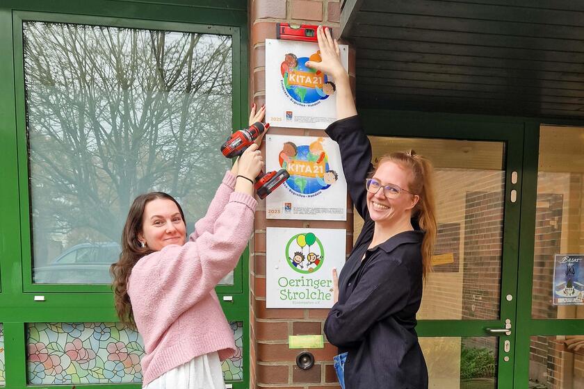 Stolz bringen Auszubildende Celina Schneider und Mitarbeiterin Jennifer Meyer das Auszeichnungsschild vor dem Eingang der Johanniter-Kita Oeringer Strolche an.