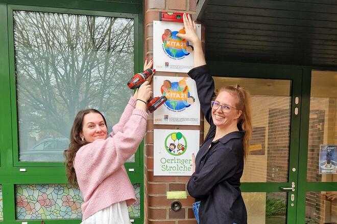 Oeringer Strolche sind Kita21 Stolz bringen Auszubildende Celina Schneider und Mitarbeiterin Jennifer Meyer das Auszeichnungsschild vor dem Eingang der Johanniter-Kita Oeringer Strolche an.