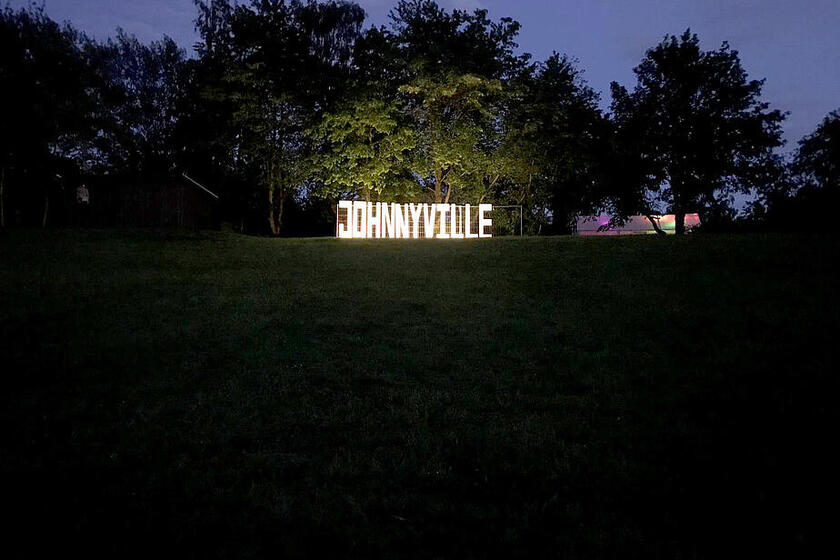 Es ist dunkel. Der Schriftzug "JohnnyVille" wird am Rande einer Wiese beleuchtet.