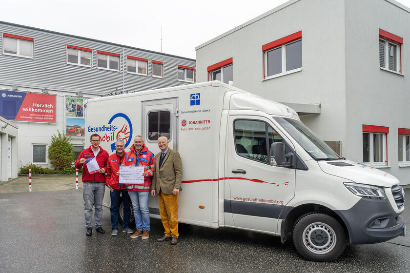 Yonathan Arnold (zuständiger Bereichsleiter), Hans-Martin-Grusnick (ehrenamtliches Mitglied des Regionalvorstandes), Thomas Müller (Mitarbeiter des Gesundheitsmobil) (v.l.) nehmen den symbolischen Scheck über 6.000 Euro von Dr. Erik Peters entgegen. Peters übergibt die Spende stellvertretend für die Schleswig-Holsteinischen Johanniter-Hilfsgemeinschaften des Johanniterordens. Sie stehen an der Dienststelle der Johanniter vor dem Gesundheitsmobil.