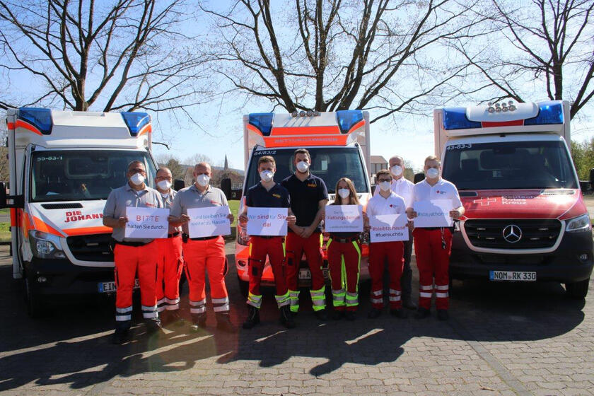 Mitarbeiter der Rettungsdienste stehen vor ihren Fahrezugen und halten Schilder hoch