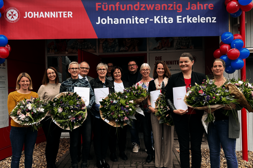 25 Jahre Johanniter-Kita Erkelenz Zwölf Frauen und Männer stehen vor dem mit Luftballons geschmückten Eingang einer Kindertagesstätte. Einige halten Blumensträuße in den Händen.