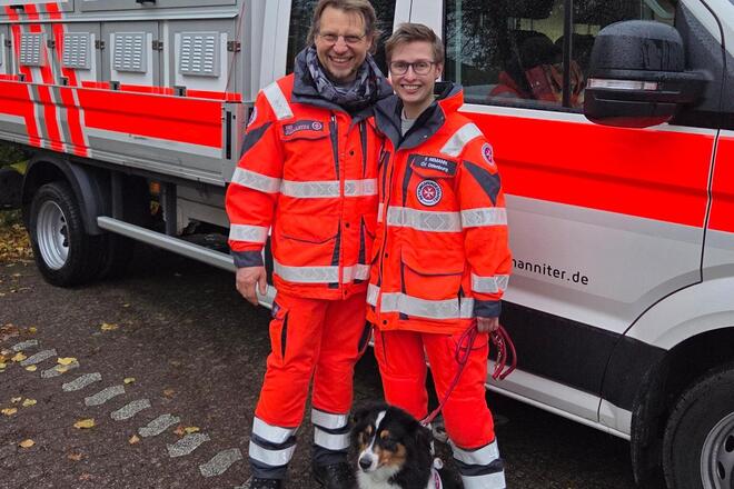 Ein Mann und eine Frau in Einsatzkleidung stehen zusammen mit einem Hund mit Kenndecke vor einem Einsatzfahrzeug.