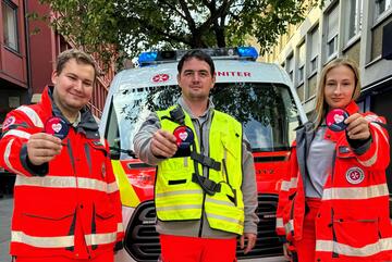 Drei Sanitäterinnen und Sanitäter der Johanniter, stehen in Einsatzkleidung vor einem Johanniter-Einsatzwagen und halte ein rundes Patch in die Kamera. Das Motiv des Patches zeigt die Zusammenarbeit zwischen den Johannitern und den Weindorf.