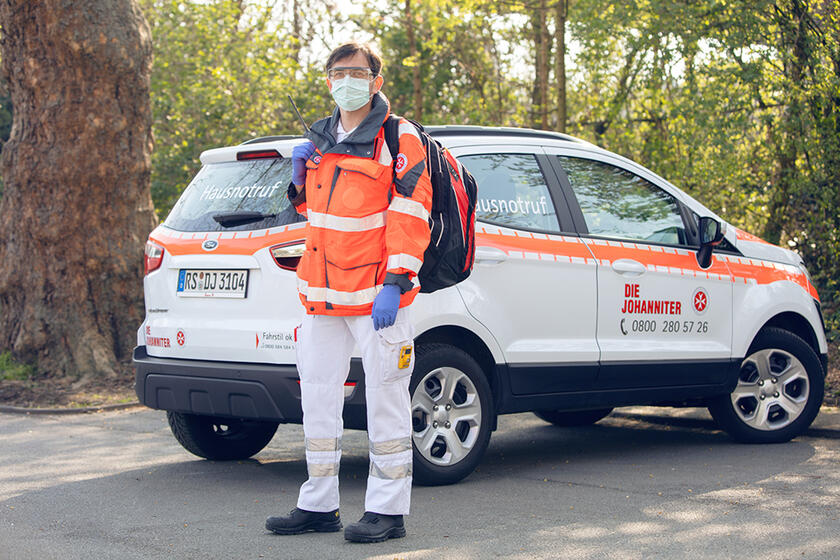 Johanniter-Mitarbeiter in Einsatzkleidung, Mundschutz und Handschuhen vor seinem Fahrzeug.