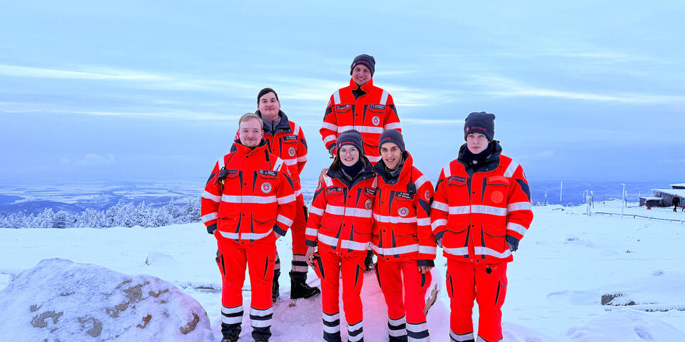 6 Johanniter und Johanniterinnen, v4 davon mit Mütze, stehen auf einem verschneiten Gipfel. 