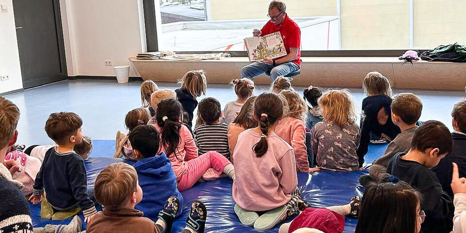 Ein Mann hält ein Buch und sitzt vor einer Gruppe von Kindern und Erwachsenen