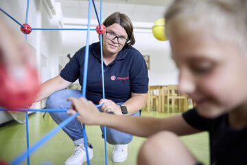 Eine Betreuerin der Johanniter beaufsichtigt ein spielerisches Konstruktionsprojekt in einer offenen Ganztagesschule. Ein Kind im Vordergrund konzentriert sich auf das Spiel.