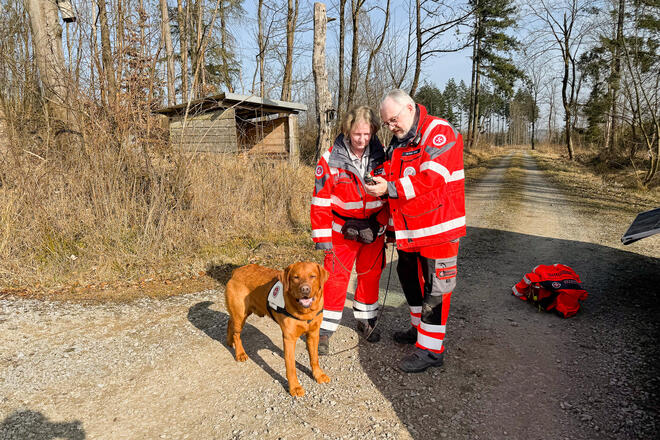 Eine Johanniterin mit einem hellbraunen Hund an der Leine, schaut gemeinsam mit einem Johanniter auf ein GPS-Gerät. Sie befinden sich in einem Wald.