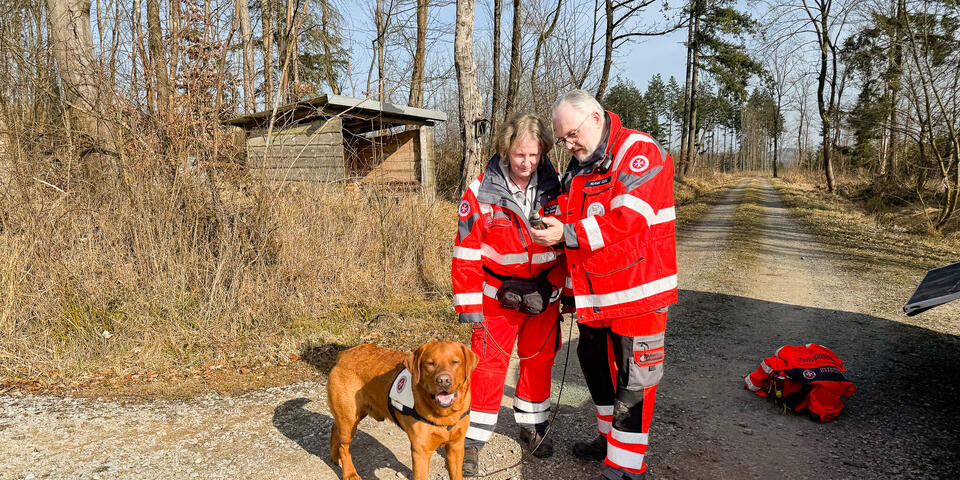 Eine Johanniterin mit einem hellbraunen Hund an der Leine, schaut gemeinsam mit einem Johanniter auf ein GPS-Gerät. Sie befinden sich in einem Wald.