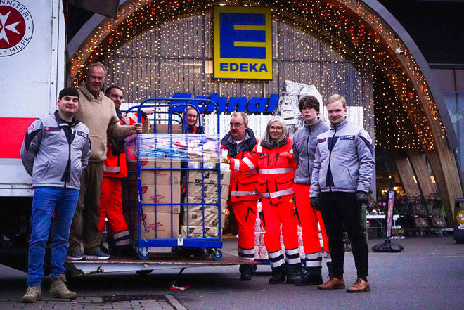 Sechs Männer und zwei Frauen, teilweise in Johanniter-Kleidung an einem LKW dessen Rampe heruntergelassen ist. Auf der Rampe steht ein gefüllter Rollbehälter.