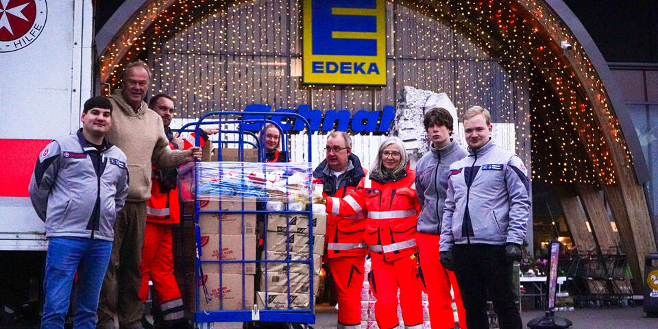 Sechs Männer und zwei Frauen, teilweise in Johanniter-Kleidung an einem LKW dessen Rampe heruntergelassen ist. Auf der Rampe steht ein gefüllter Rollbehälter.
