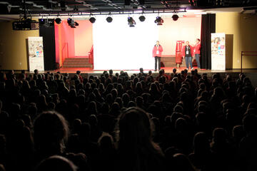 Eine Aula mit vielen Menschen, die Bühne vorne ist beleuchtet und Personen in Johanniter-Jugend-Kleidung reden.