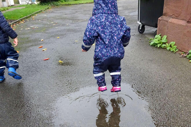 Kita-Kind springt mit Regenklamotten und Gummistiefeln in eine Pfütze. 