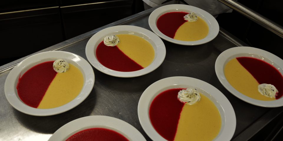 Ein Tablettwagen, auf dem sechs Teller mit Suppe stehen. Die Suppe ist zur Hälfte rot und zur anderen Hälfte hellgelb