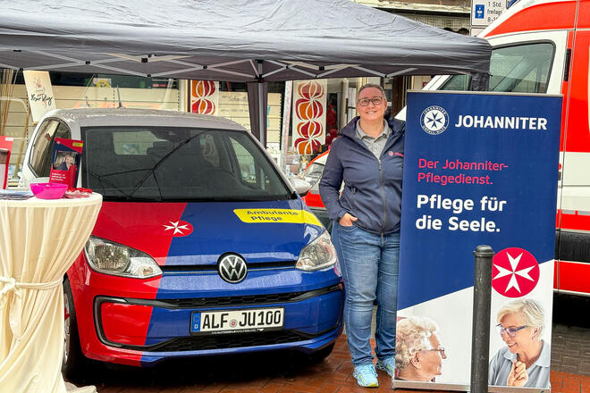 Eine Johanniterin unter einem Pavillon neben einem Roll-Up mit Pflegemotiv. Links neben ihr ein Johanniter-Auto