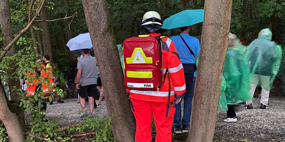 Ein Johanniter im Wald. Es regnet. Hinter ihm sind Personen mit Schirmen und Regenjacken zu sehen.