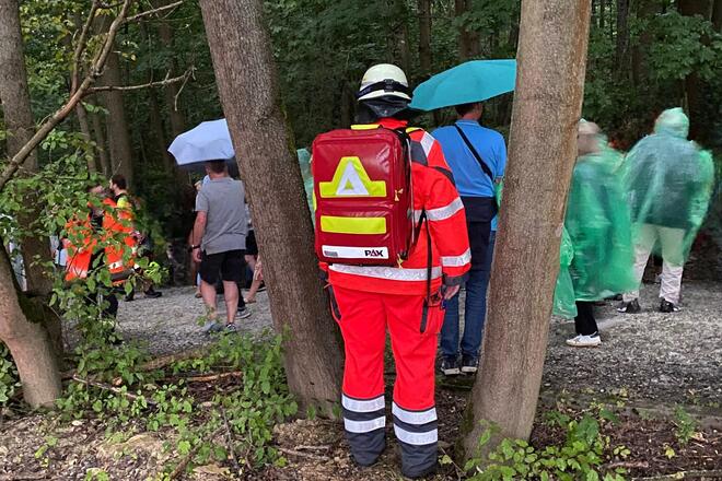 Ein Johanniter im Wald. Es regnet. Hinter ihm sind Personen mit Schirmen und Regenjacken zu sehen.