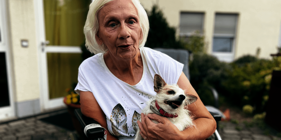 Eine weißhaarige Frau im weißen T-Shirt sitzt in einem Rollstuhl und hält einen kleine Hund auf dem Schoß.