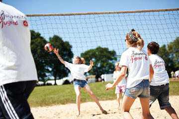 Jugendliche der Johanniter-Jugend spielen bei sonnigem Wetter Volleyball auf einem Sandplatz. Alle tragen weiße T-Shirts mit dem Logo der Johanniter-Jugend.