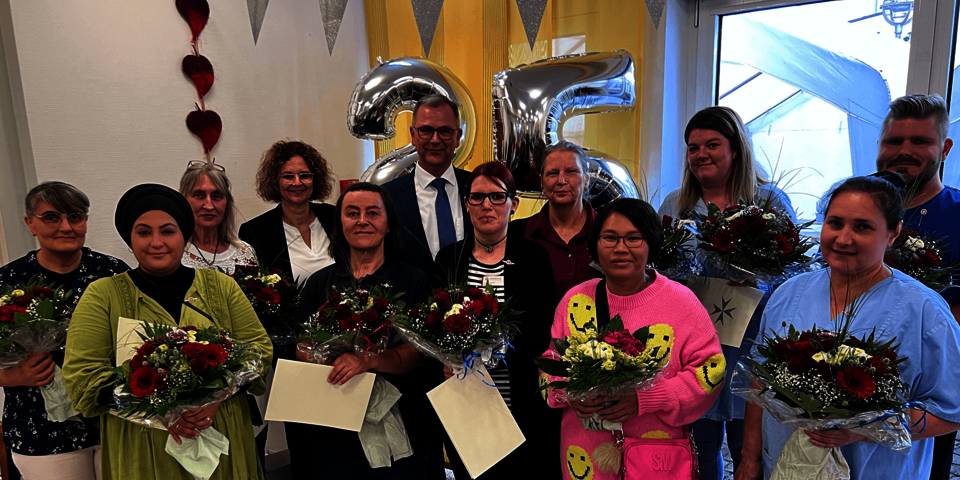 Eine Gruppe von 12 Menschen vor einer silbernen 25 als Luftballons; die meisten halten Blumensträuße in den Händen