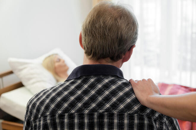 Ein Mann sitzt vor einem Bett, in dem eine Frau liegt. Eine Person außerhalb des Bildes hat ihre Hand auf seine Schulter gelegt.