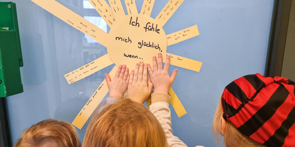 Kinder fassen eine Sonne aus Pappe an. Auf der Sonne steht "Was mich glücklich macht"