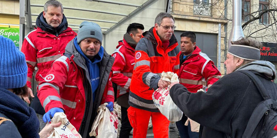 Mehrere Johanniter stehen auf der Laderampe eines LKW und verteilen Baumwolltaschen an Obdachlose und Bedürftige zur Johanniter-Suppenküche. 