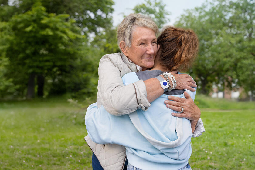 Zwei Frauen umarmen sich. Die ältere Frau trägt einen Handsender am Handgelenk. Im Hintergrund ist ein Park zu sehen.