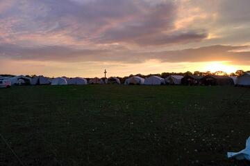 Eine Wiese beim Sonnenuntergang. Am Rand der Wiese stehen Zelte und es ist ein großes Kreuz im Gegenlicht zu sehen.