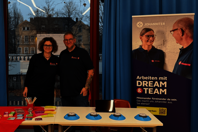 Zwei Johanniter-Mitarbeitende stehen zusammen hinter einem Infostand auf einer Messe. Der Stand ist dekoriert mit Flyern, einer roten Johanniter-Tasse und einem weißen Roll-Up mit dem Text "Arbeiten mit DREAM & TEAM".