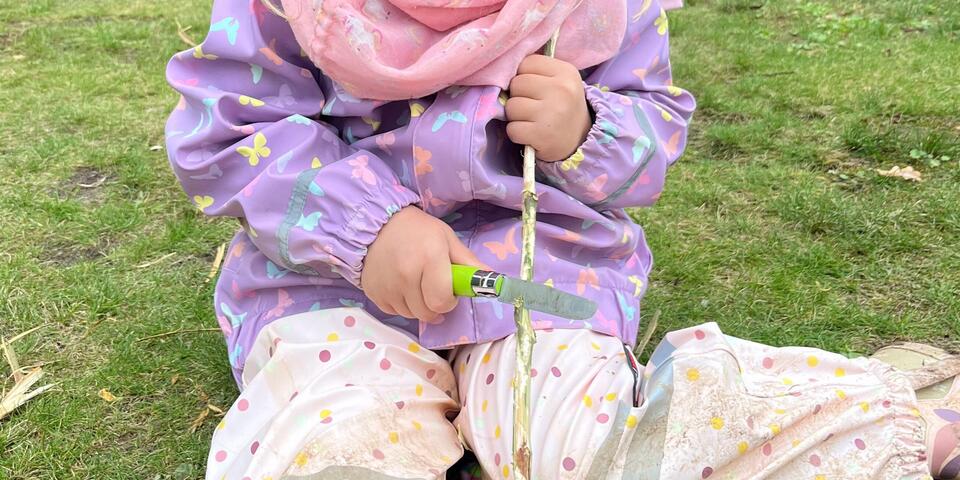 Ein Kind schnitzt einen Stock und sitzt dabei auf einem Rasen.