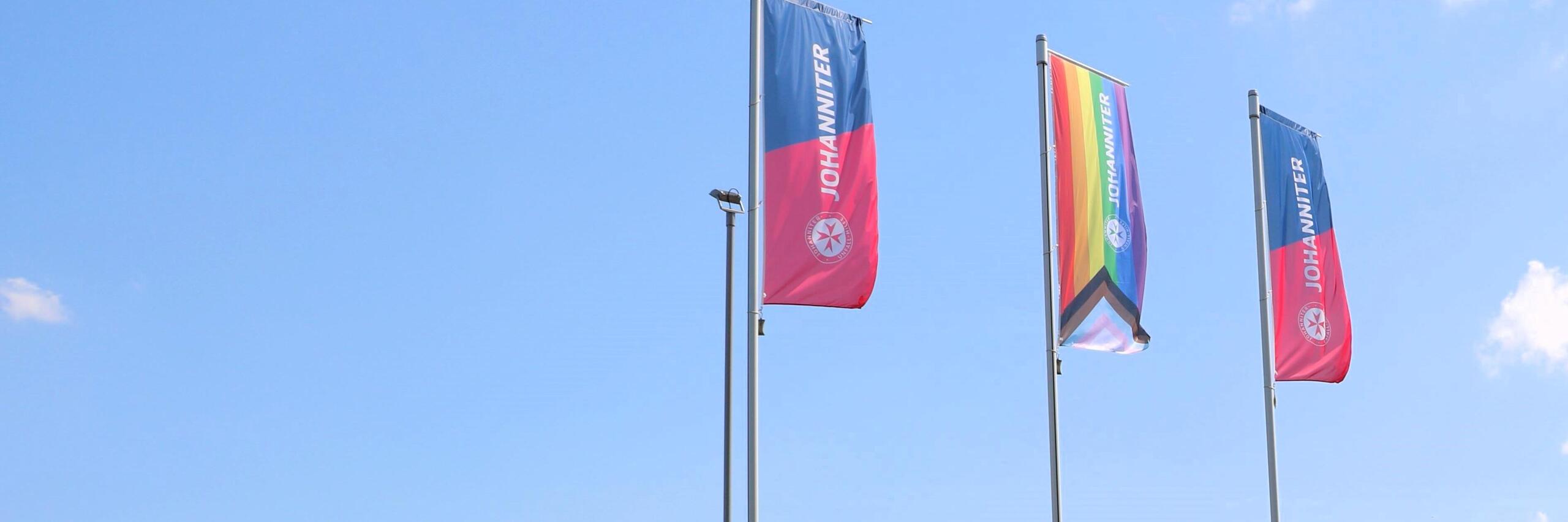 Das Bild zeigt drei Johanniter-Fahnen vor der Regionalgeschäftsstelle in Blomberg. Die beiden äußeren in den Johanniter-Farben Rot und Blau, die mittlere Fahne ist in Regenbogenfarben.