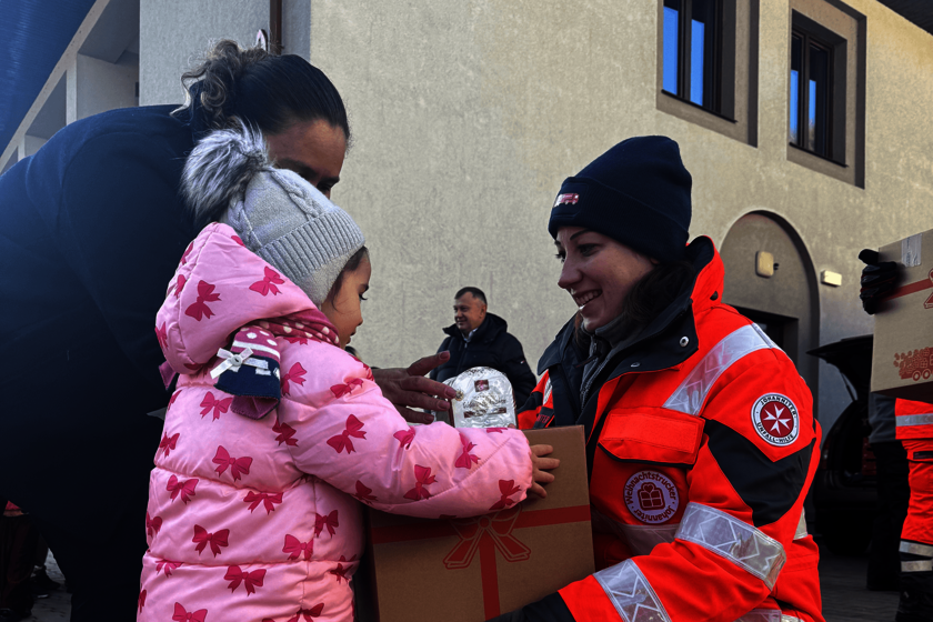 Eine Johanniter-Helferin in orangeroter Einsatzkleidung übergibt draußen ein Paket mit Geschenkaufdruck an ein kleines Mädchen im rosafarbenen Wintermantel. 