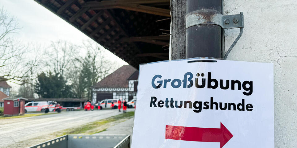 Ein Schild auf dem "Großübung - Rettungshunde" steht an einem Abflussrohr einer Regenrinne an einem weißen Gebäude.