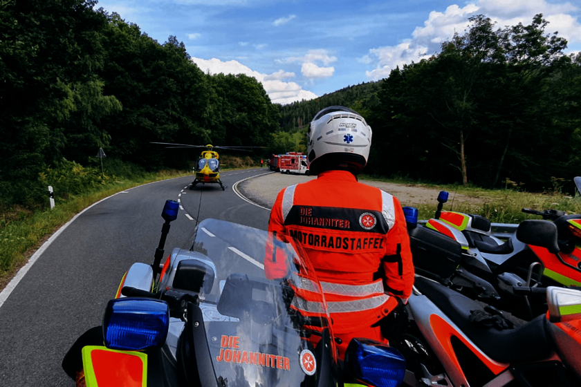 Ein Einsatzmotorrad und ein Fahrer in Einsatzkleidung und mit Helm sind von hinten zu sehen.  Weiter vorne auf der Landstraße sind ein Rettungshubschrauber und ein Rettungswagen zu sehen.
