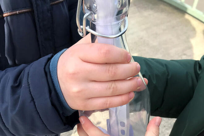 Man sieht die Hände von zwei Kita-Kindern, wie sie eine Flasche, in der sich eine Nachricht befindet, festhalten.