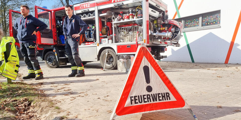 Zwei Feuerehrleute vor einem Feuerwehrauto. Im Vordergrund ein Warnschild auf dem "Feuerwehr" steht