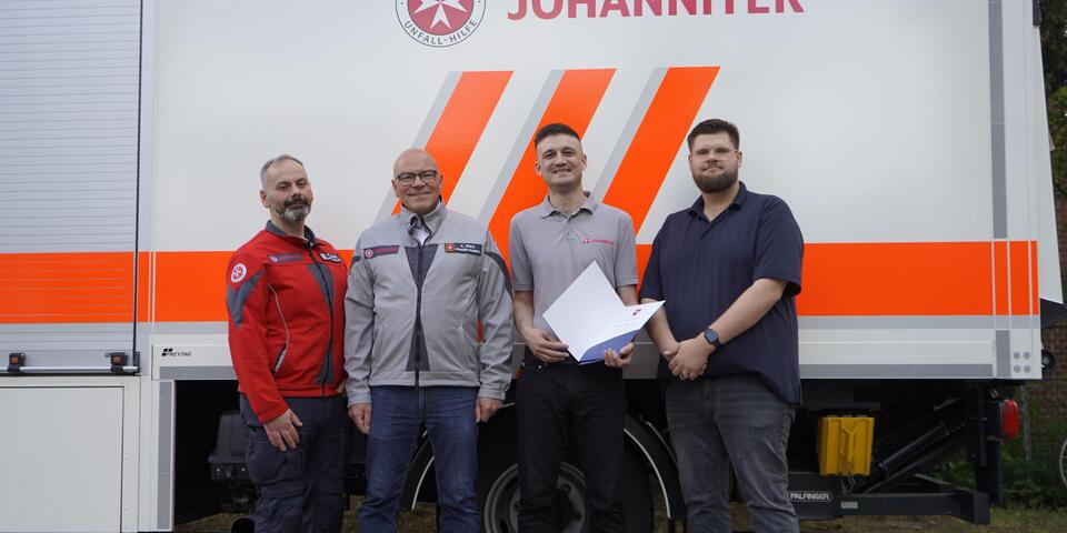 Vier Männer vor einem Johanniter-Fahrzeug. Einer hält ein Zertifikat
