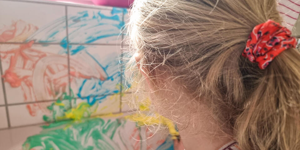Ein kleines Mädchen malt mit einem Pinsel bunte Farben an weiße Fliesen